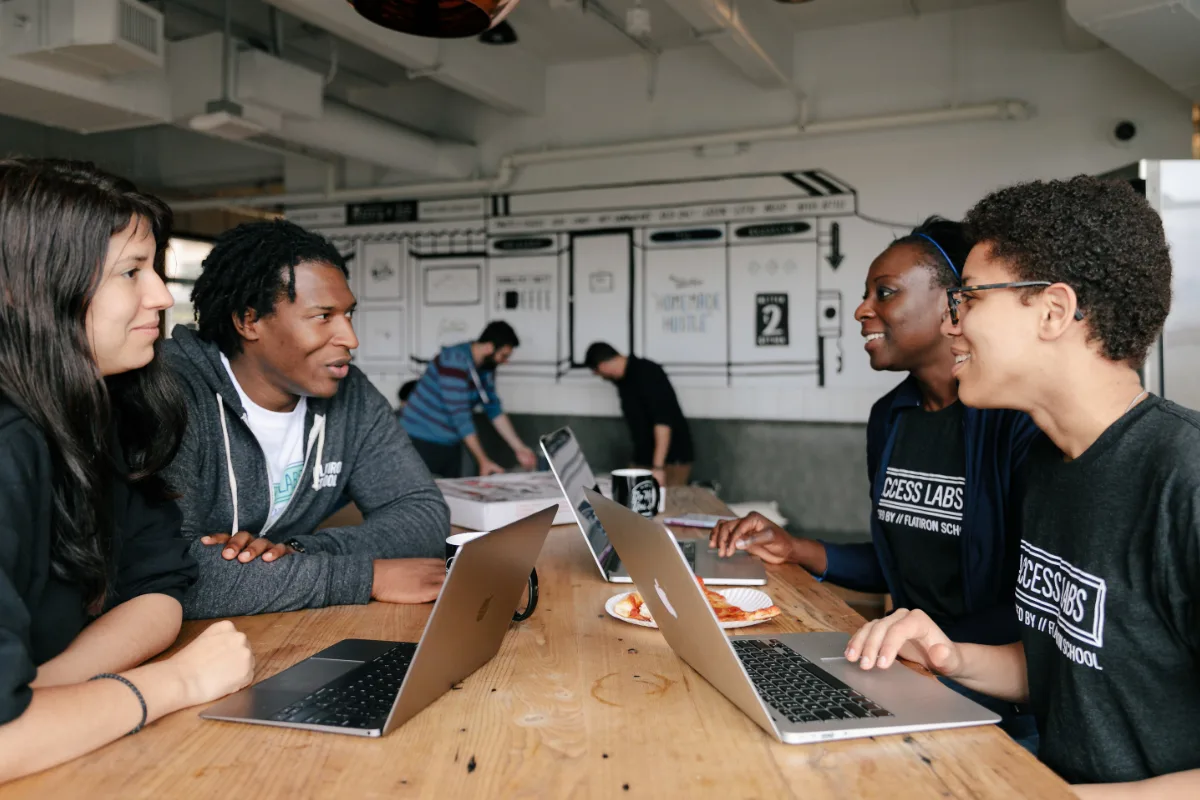 Four young individuals engaged in a discussion around a table with laptops and food, in a modern workspace.